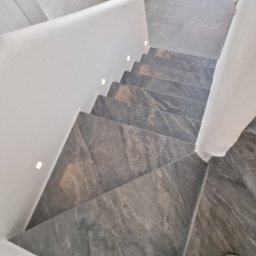 Home Future Chojnowski Andrzej - Nowoczesne schody z szarego kamienia z subtelnym oświetleniem LED wzdłuż białej ściany. Widok z góry, fragment butów w kadrze.