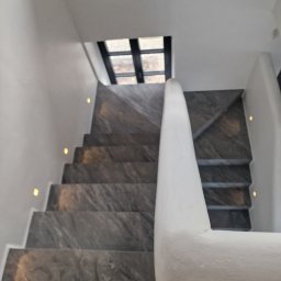 Home Future Chojnowski Andrzej - Schody wykończone szarym kamieniem z oświetleniem punktowym wzdłuż białej ściany i balustrady, widok z góry na dół, okno w tle.