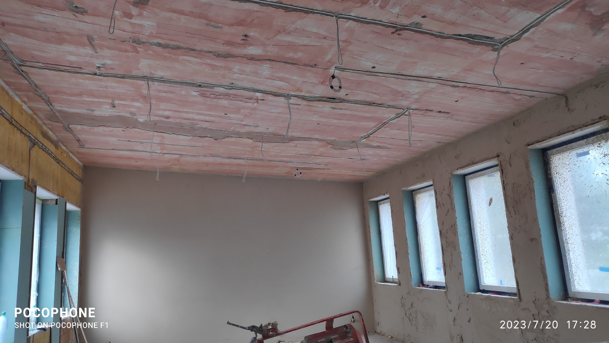 Surowy stan wnętrza z instalacją elektryczną pod tynk na suficie, ściany przygotowane pod gładzie, widoczne okna z folią ochronną i sprzęt budowlany na podłodze.