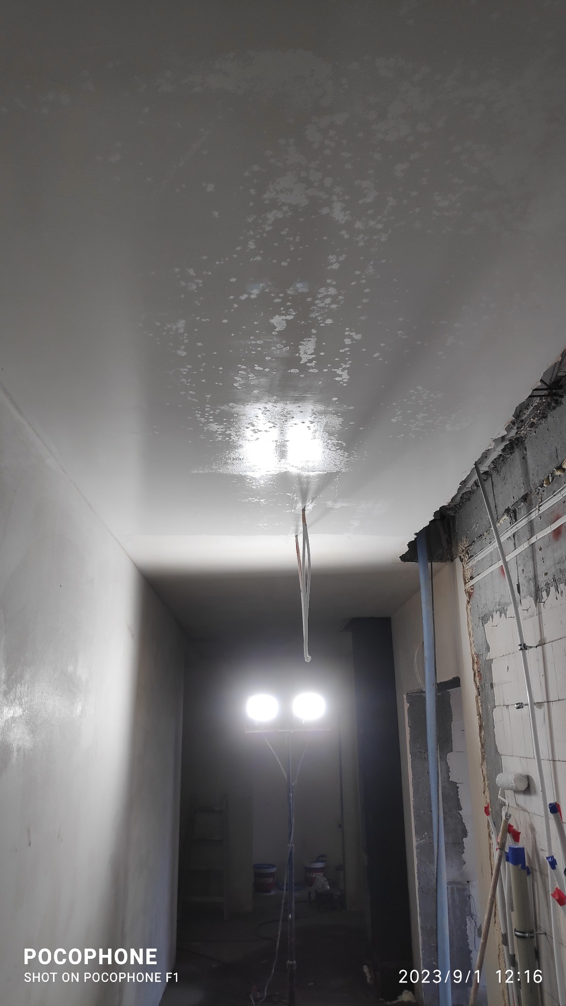 Niedokończony sufit z widocznymi plamami po nałożeniu pierwszej warstwy, oświetlony lampą budowlaną, fragment ściany w stanie surowym z instalacjami.