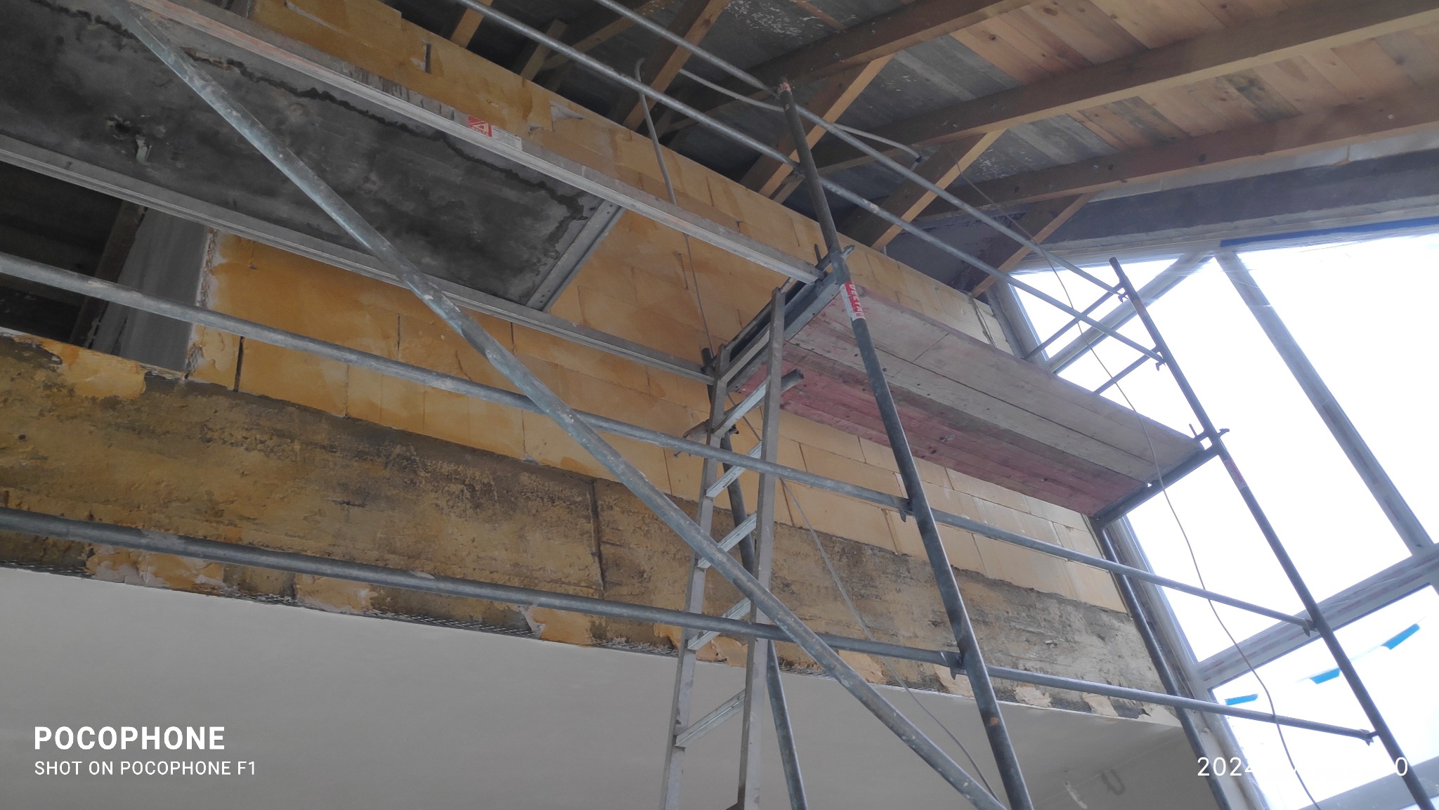 Wnętrze w trakcie prac wykończeniowych: widok z dołu na rusztowanie, fragment ściany z cegieł, surowy strop drewniany i okno dachowe w trakcie montażu.