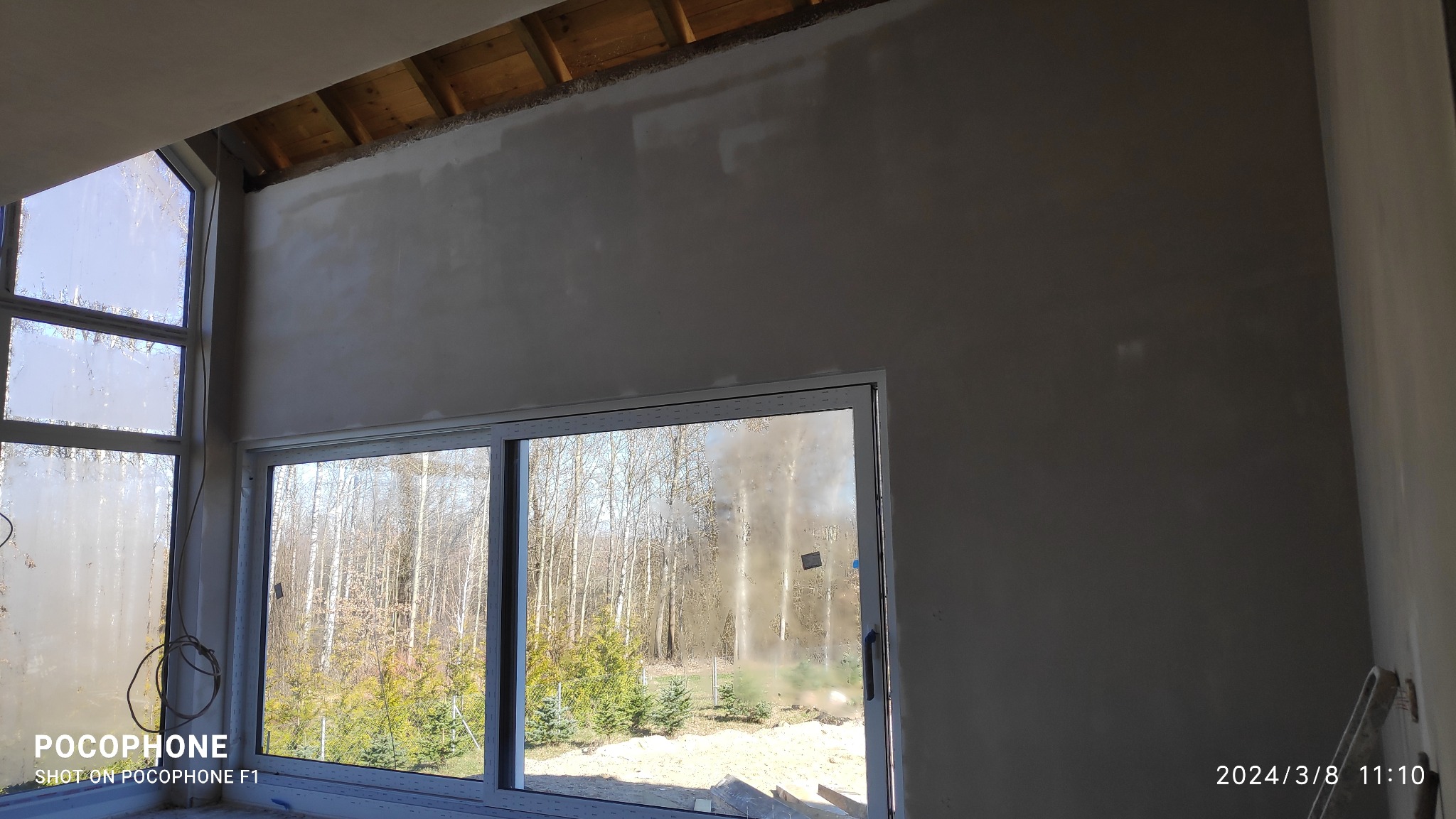 Surowa ściana wewnętrzna po szpachlowaniu, widoczne drewniane elementy konstrukcyjne stropu, okna z widokiem na zewnątrz, w tle las.