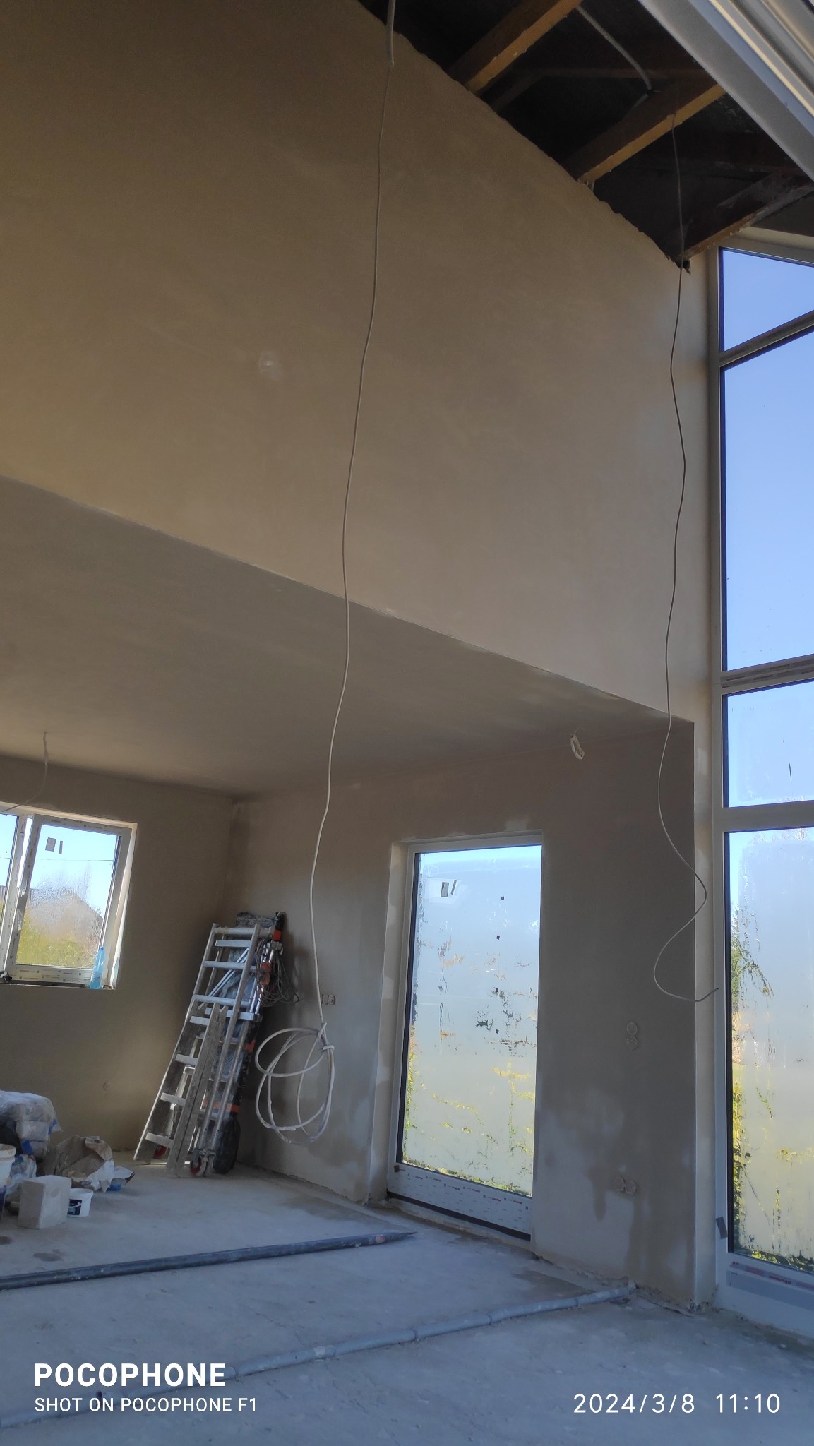 Wysokie wnętrze w trakcie prac wykończeniowych, ściany pokryte świeżym tynkiem, widoczne okna, drabina i wystające kable elektryczne.