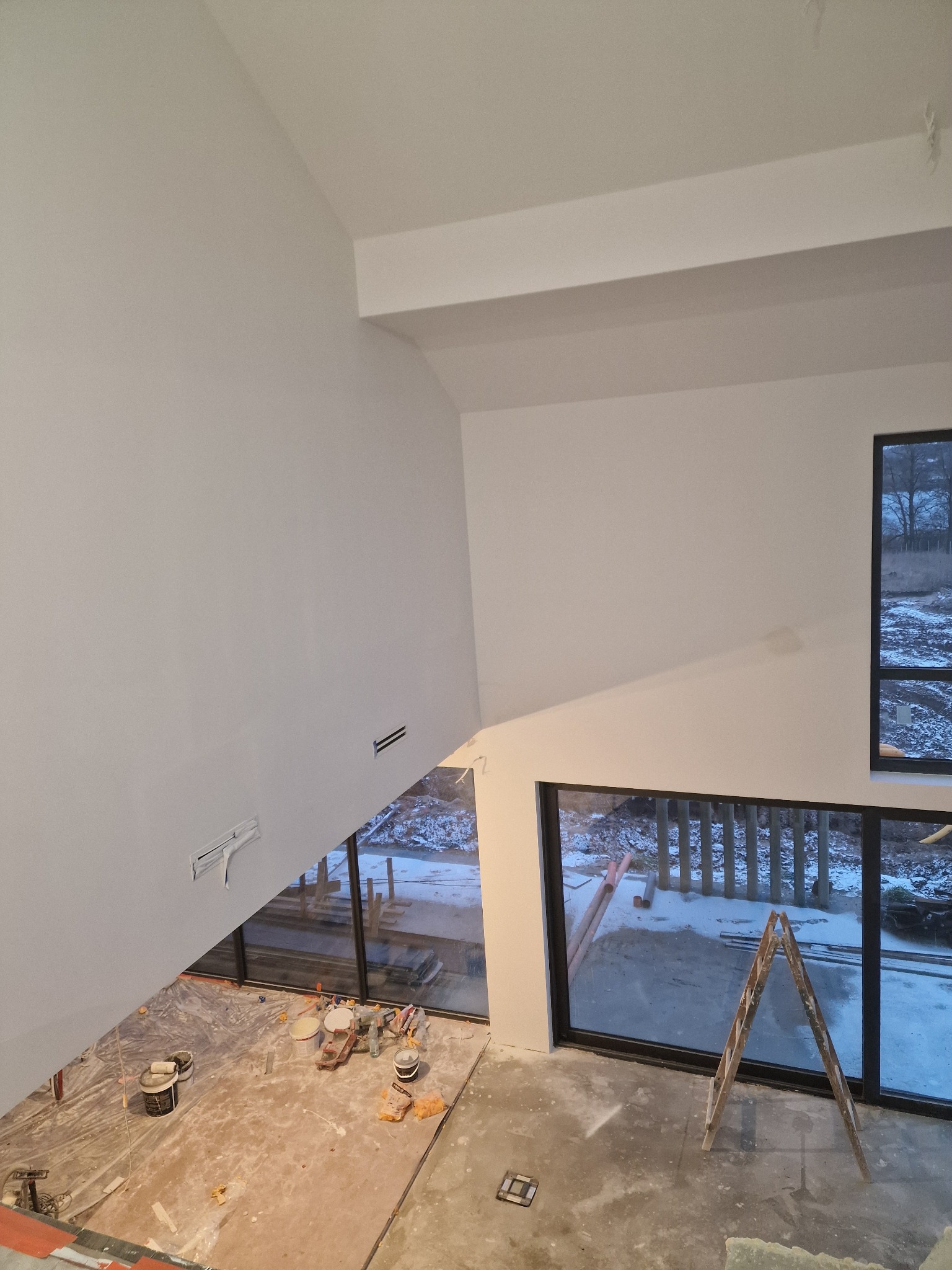 Wnętrze w trakcie prac wykończeniowych: świeżo pomalowane białe ściany w wysokim pomieszczeniu z dużymi oknami, drabina oparta o okno, widoczne puszki z farbą i narzędzia na podłodze zabezpieczonej...