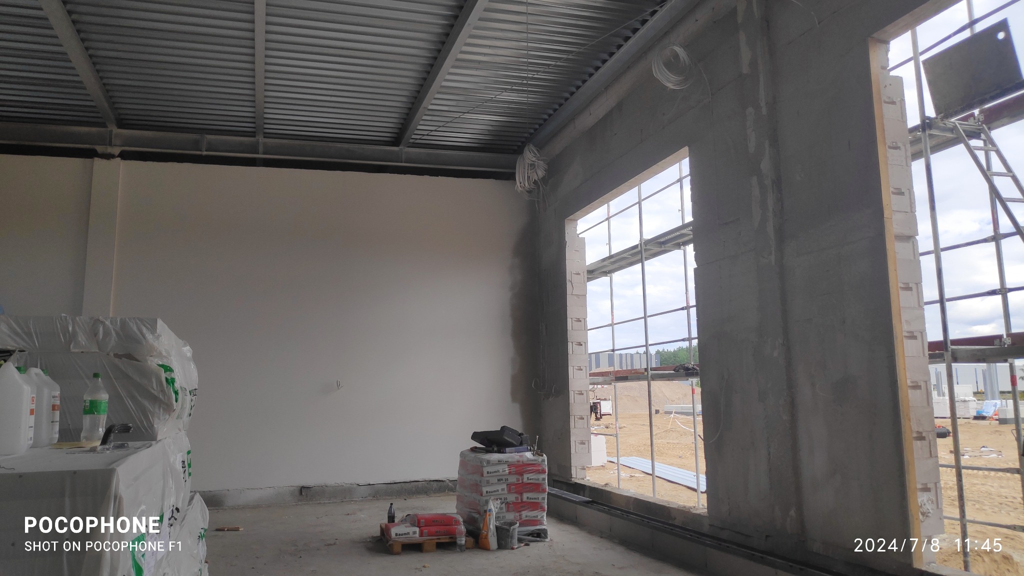 Wnętrze w trakcie prac budowlanych: ściana po obróbce, palety z zaprawą, widoczne rusztowania i surowa konstrukcja dachu.