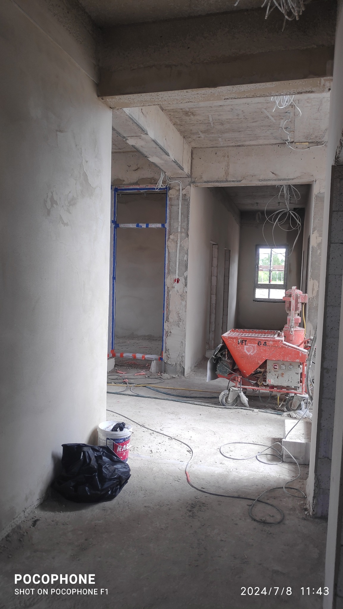 Wnętrze w stanie surowym z widoczną maszyną do tynkowania PFT G4, wiadro z zaprawą i workiem na podłodze, ściany pokryte szarym tynkiem, widoczne przewody elektryczne na suficie.