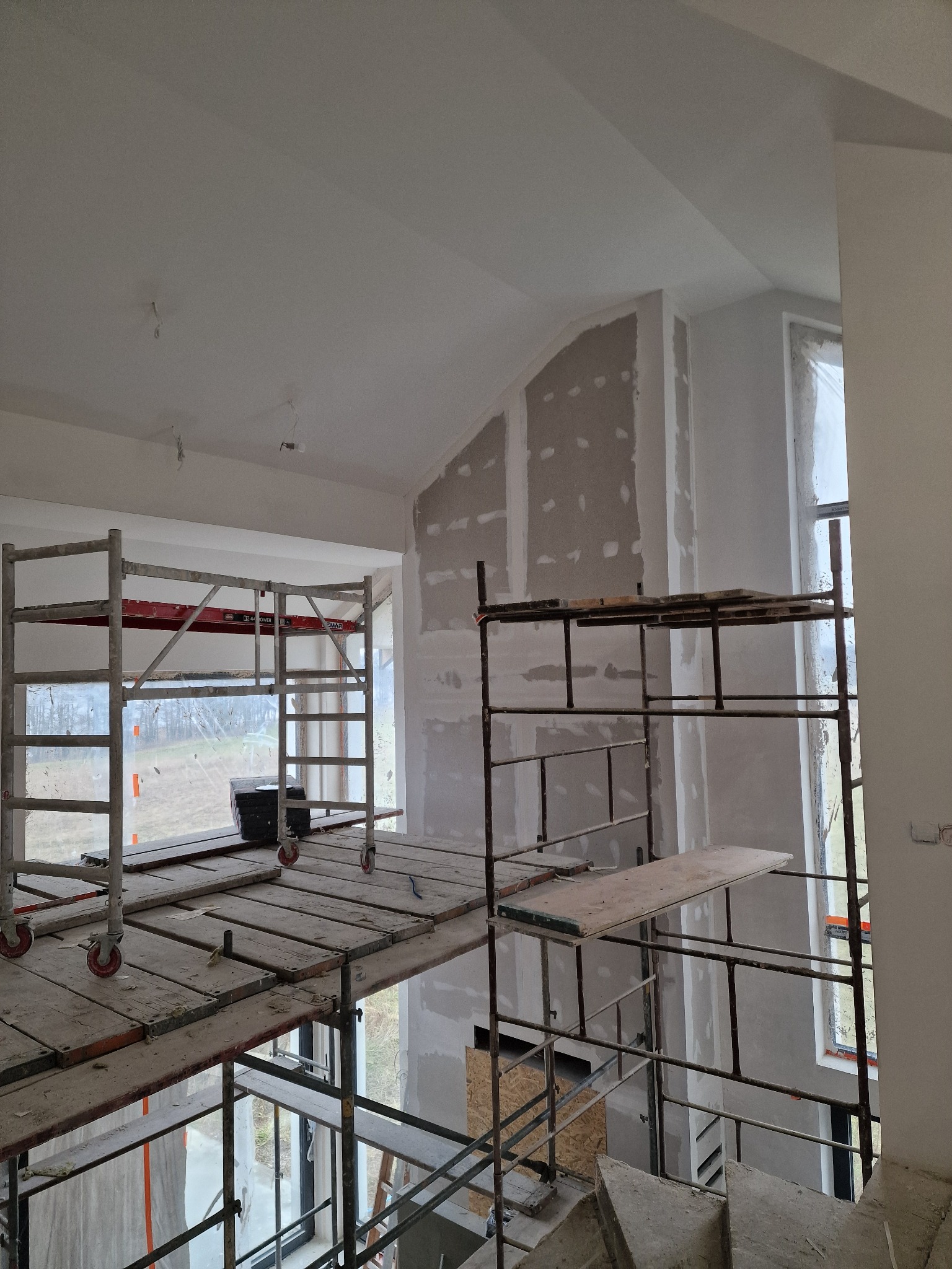 Wnętrze w trakcie prac wykończeniowych: widoczne rusztowania, ściany z fragmentami nałożonej zaprawy, okna zabezpieczone folią.