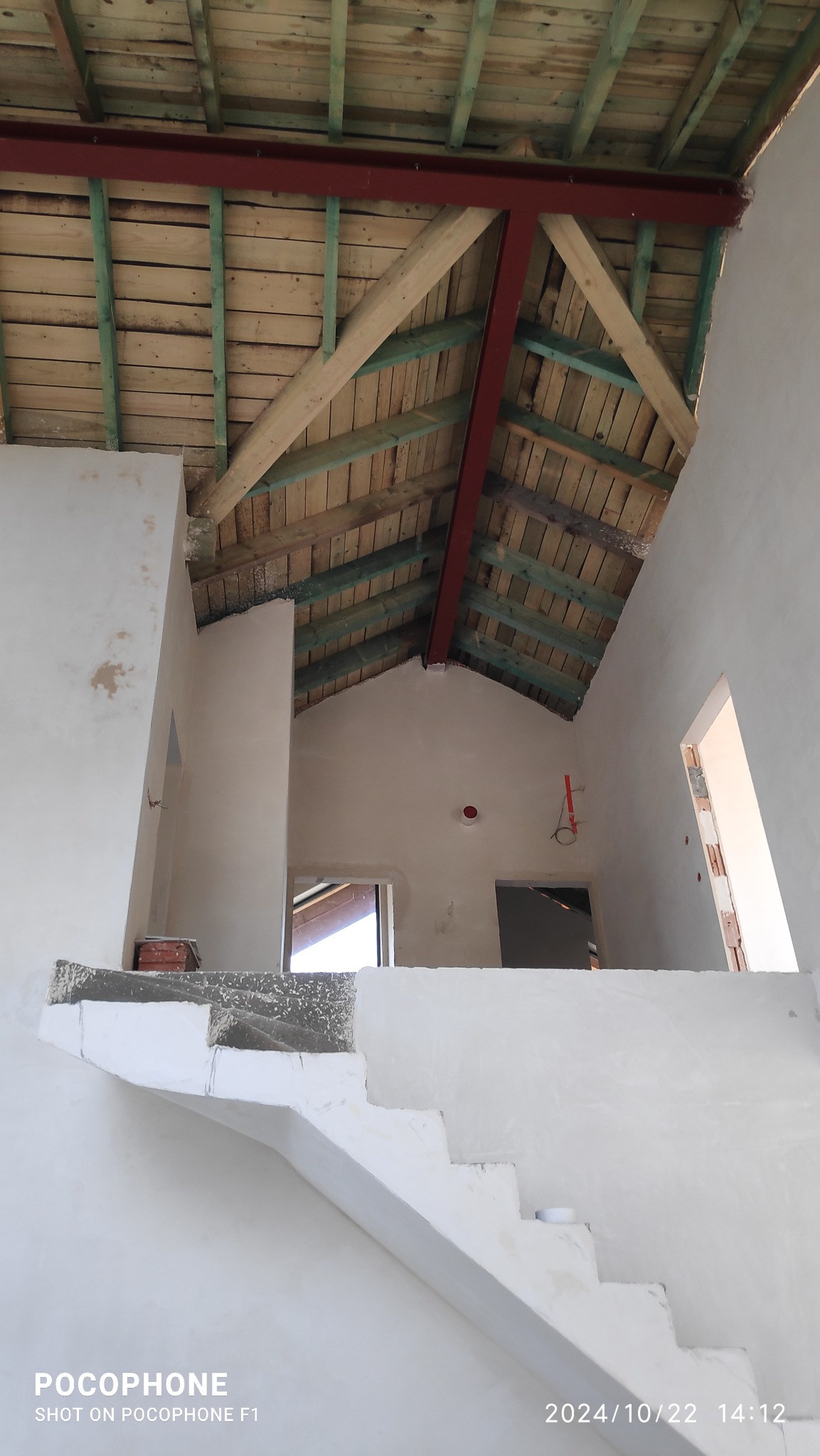 Surowe wnętrze w trakcie wykańczania: widoczne schody z surowego betonu, ściany w trakcie szpachlowania, drewniany strop z elementami stalowymi, okna bez wykończenia.