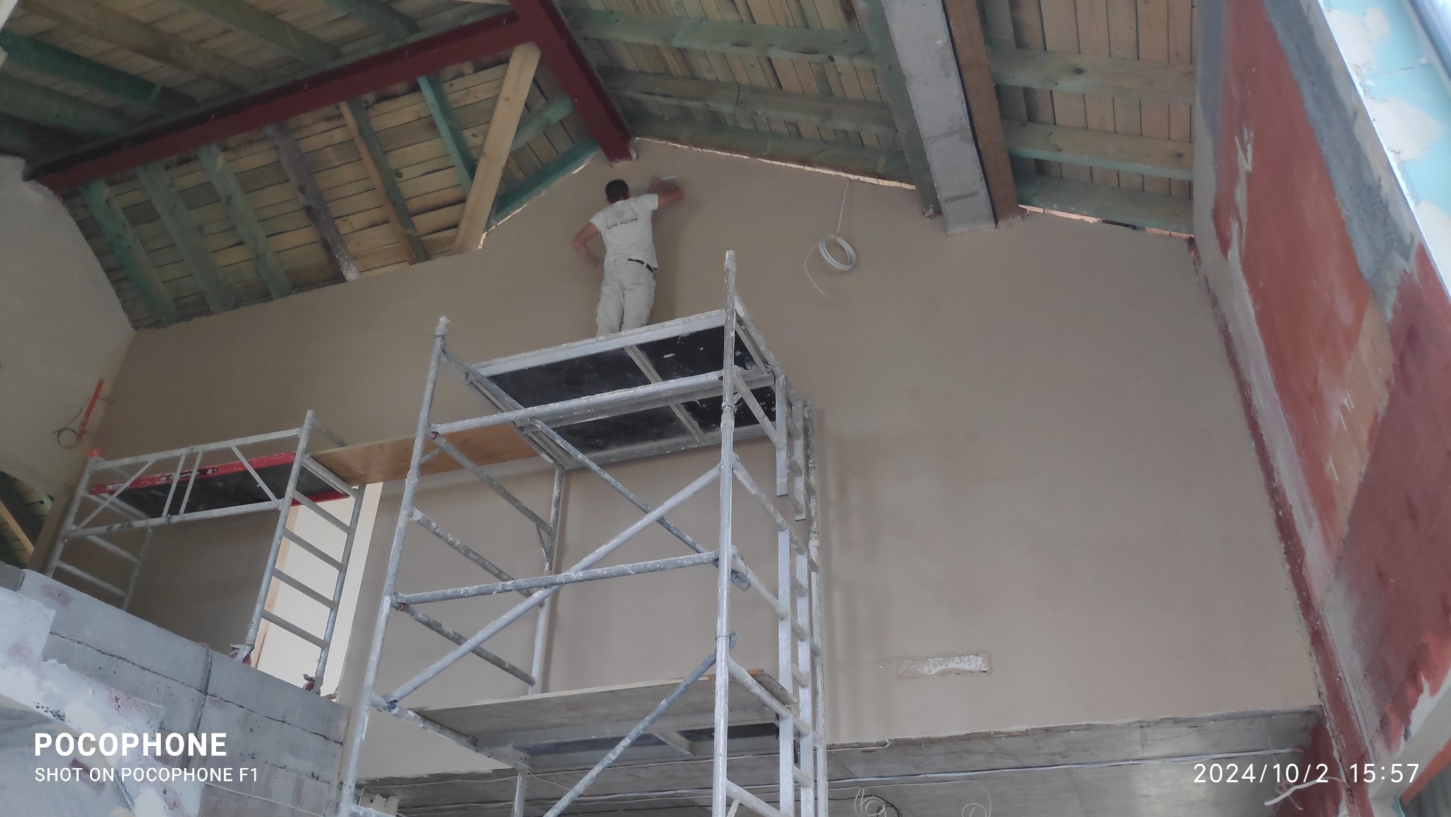 Pracownik na rusztowaniu aplikuje gładź na ścianę pod skośnym dachem, widoczne drewniane elementy konstrukcyjne, przewody elektryczne i fragmenty czerwonej ściany.