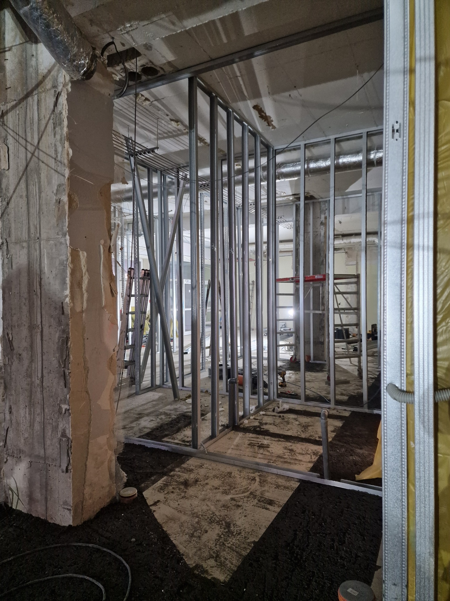 Metalowa konstrukcja ścian działowych w surowym wnętrzu, widoczne instalacje i drabina. Prace wykończeniowe w trakcie realizacji, surowy beton i elementy izolacyjne.