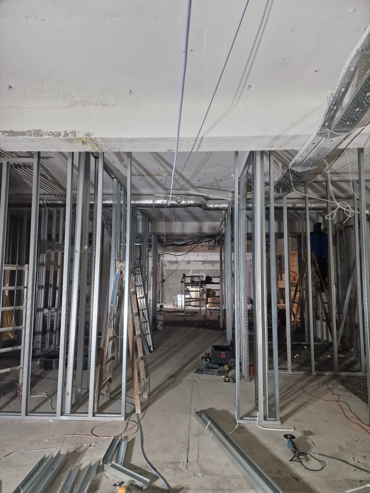 Wnętrze w trakcie budowy: metalowe profile ścian działowych, drabiny, narzędzia i instalacje pod sufitem. Widoczny pracownik na drabinie w oddali.
