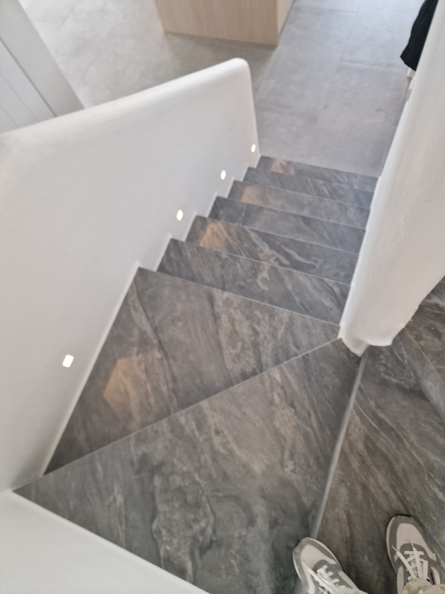 Nowoczesne schody z szarego kamienia z subtelnym oświetleniem LED wzdłuż białej ściany. Widok z góry, fragment butów w kadrze.
