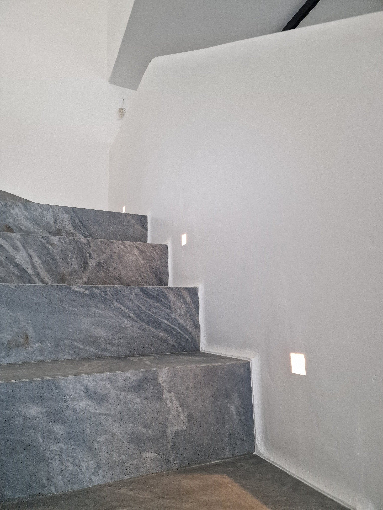 Minimalistyczne schody z szarego kamienia z subtelnym oświetleniem punktowym w białej ścianie. Nowoczesne wykończenie wnętrza z dbałością o detale.