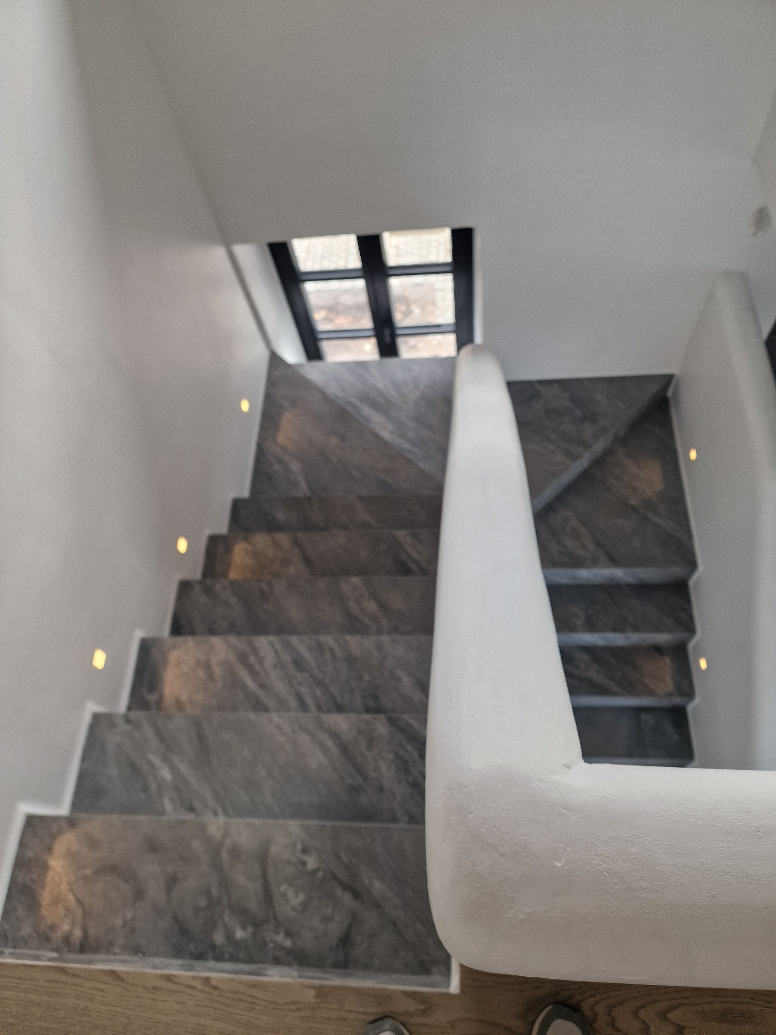 Schody wykończone szarym kamieniem z oświetleniem punktowym wzdłuż białej ściany i balustrady, widok z góry na dół, okno w tle.