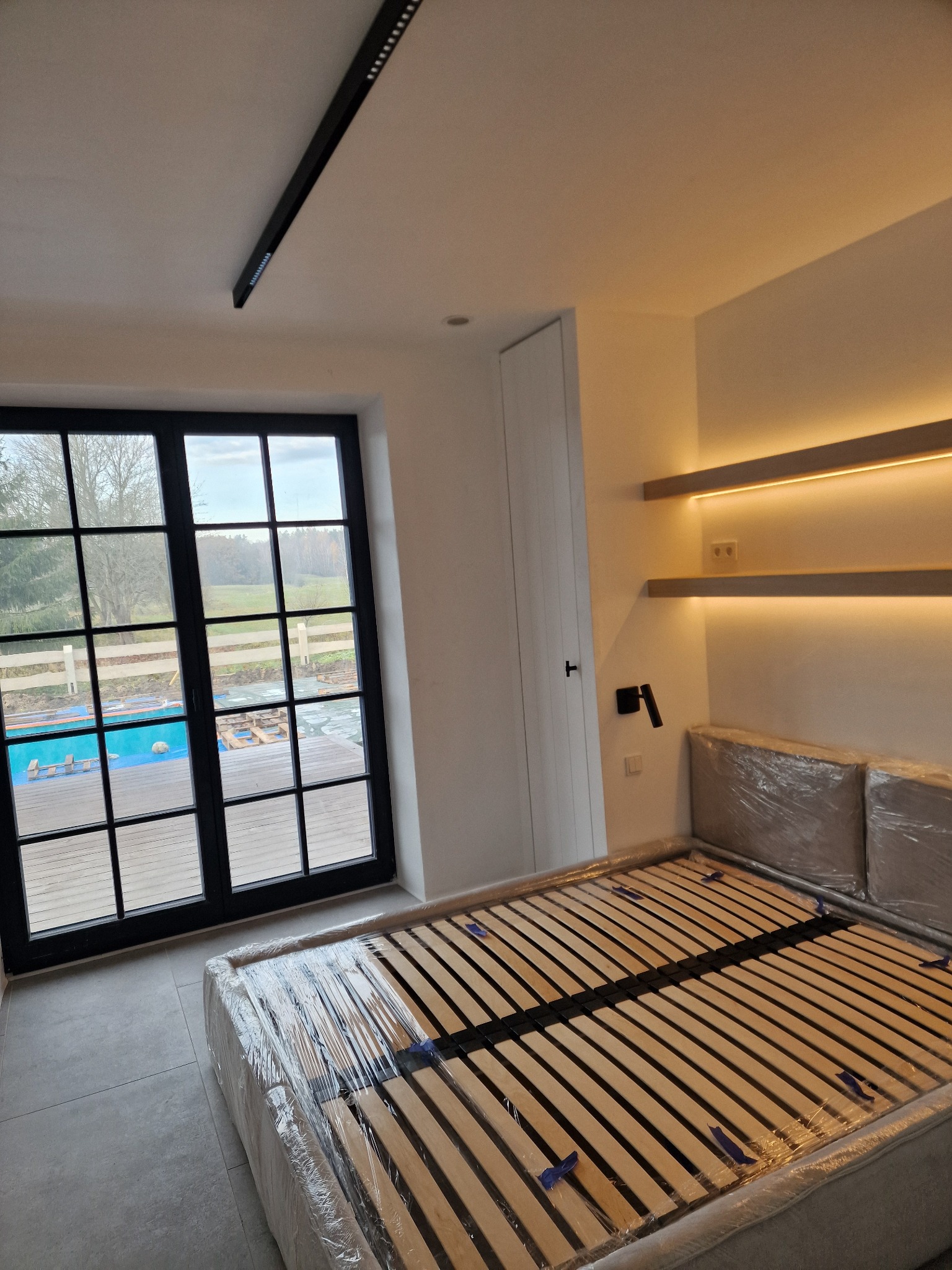 Nowoczesna sypialnia z oknem na ogród, podświetlane półki i listwa LED na suficie. Łóżko w folii ochronnej, jasne ściany i podłoga. Minimalistyczny design.