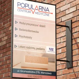 Szyld reklamowy Centrum Medycznego Popularna na ceglanej ścianie, oferujący medycynę pracy, badania kierowców, psychotesty oraz lekarza rodzinnego i pediatrę, z adresem w Warszawie.
