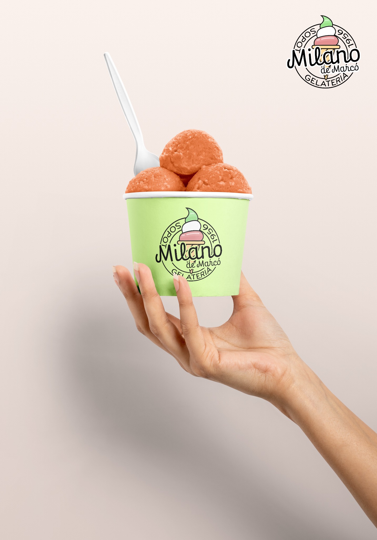 Dłoń trzymająca zielony kubek z trzema kulkami pomarańczowych lodów i białą łyżeczką, widoczne logo lodziarni Milano de Marco Gelateria.