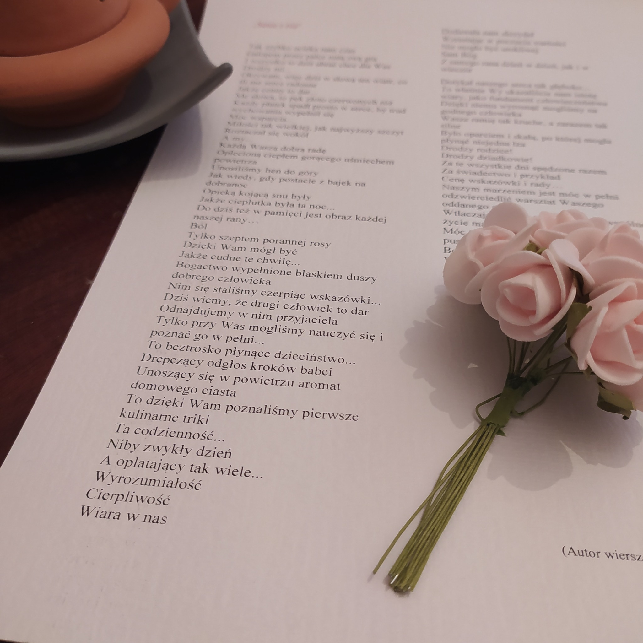 Strona z wydrukowanym wierszem, obok bukiecik różowych, sztucznych róż i filiżanka z napojem w tle.