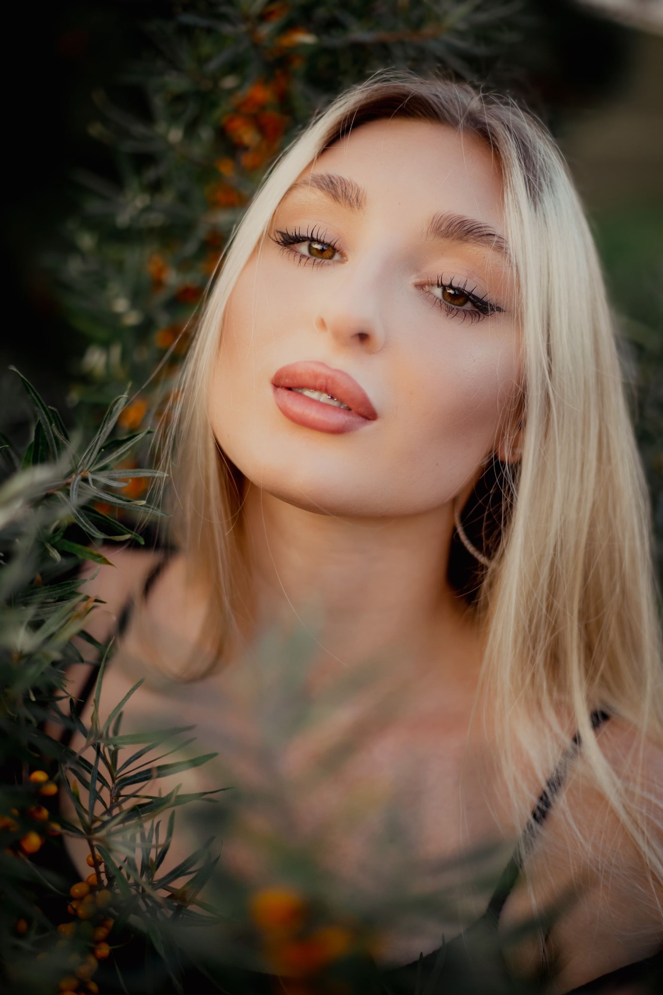 Portret młodej kobiety z blond włosami, pozującej w plenerze z gałązkami rokitnika w tle i na pierwszym planie, patrzącej w dół.