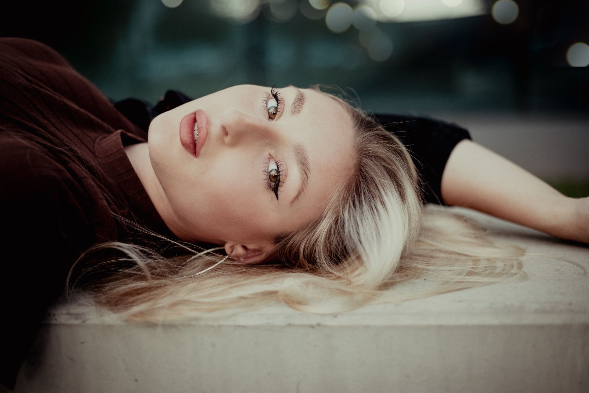 Portret leżącej blondynki z kolczykami kołami, skierowanej w górę, z widocznym makijażem oczu i ust oraz brązową bluzką.