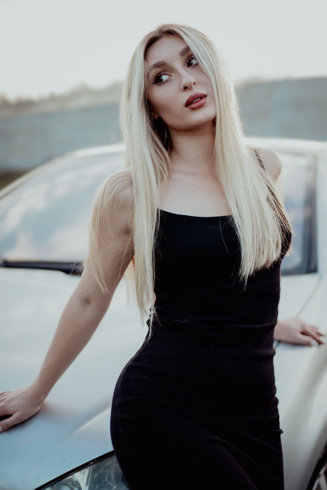 Portret młodej kobiety z długimi blond włosami, ubranej w czarną sukienkę, opierającej się o srebrny samochód i spoglądającej w bok.