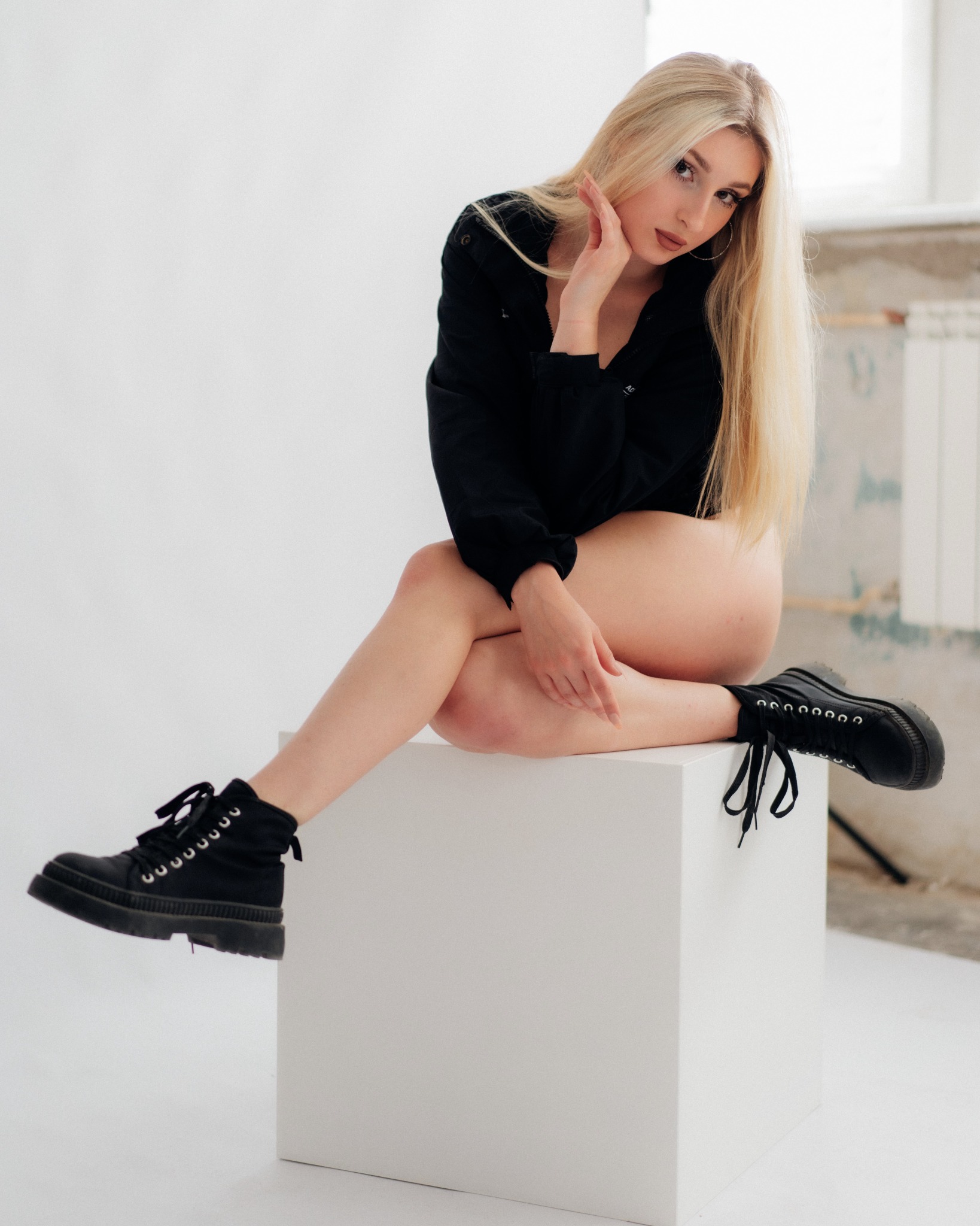 Portret blondynki w czarnej bluzce i masywnych butach, siedzącej na białym sześcianie w minimalistycznym studiu.