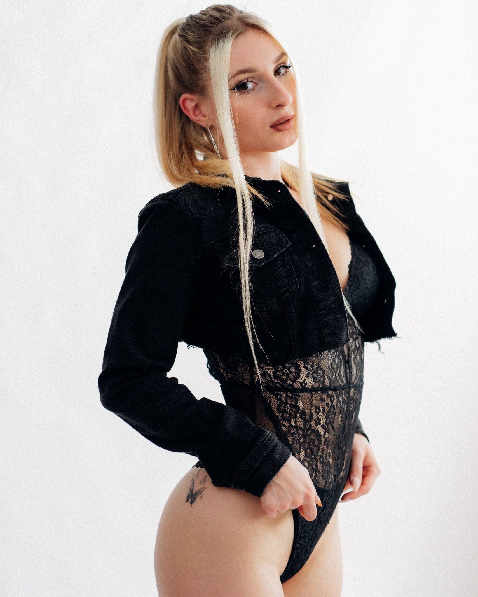 Portret kobiety w czarnej koronkowej bieliźnie i jeansowej kurtce w Rokietnicy, z blond włosami spiętymi w kucyk i subtelnym tatuażem motyla na biodrze, pozującej na jasnym tle.