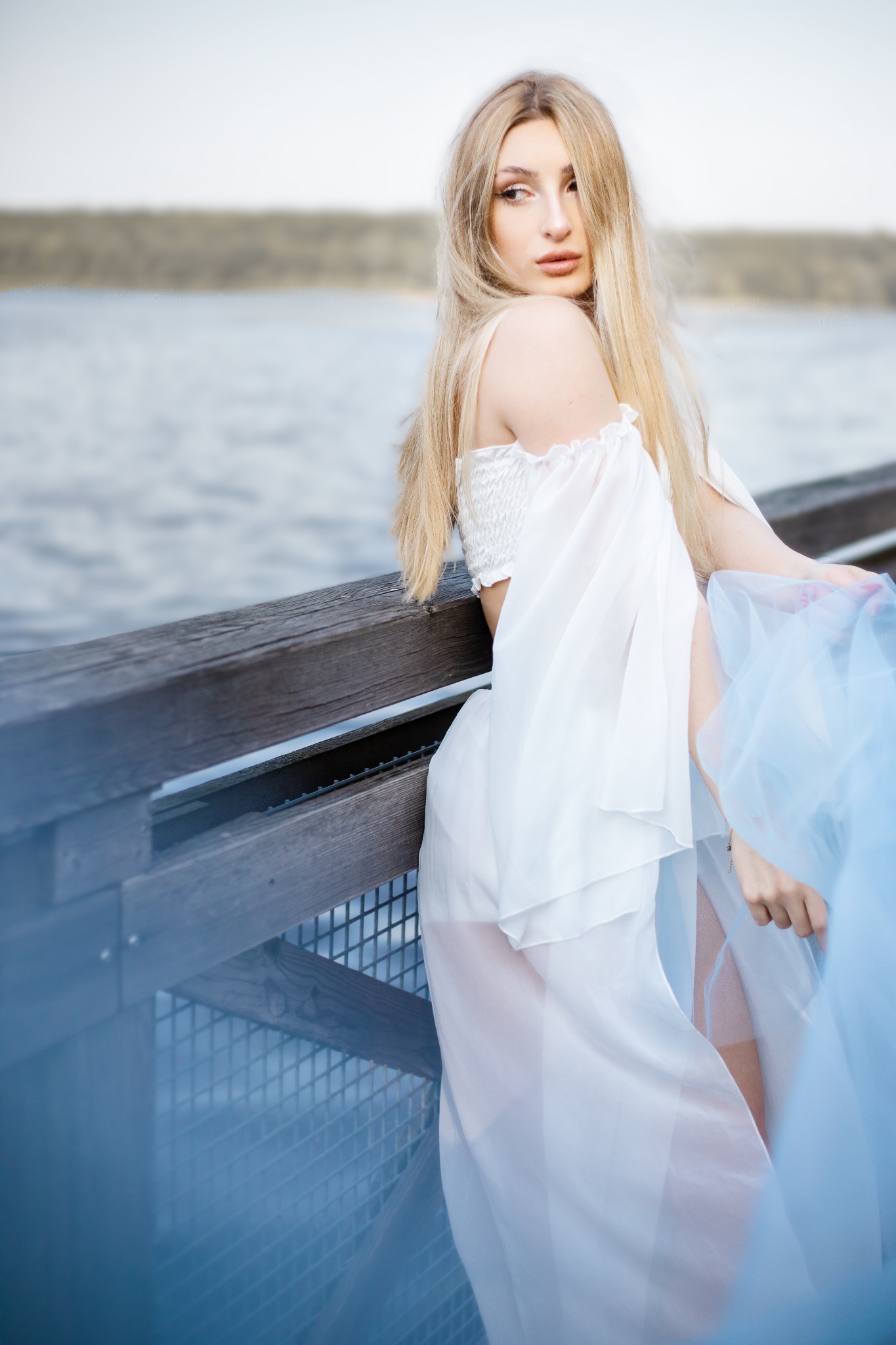 Blondwłosa kobieta w zwiewnej białej sukience pozuje na drewnianym pomoście nad wodą, trzymając błękitny materiał.