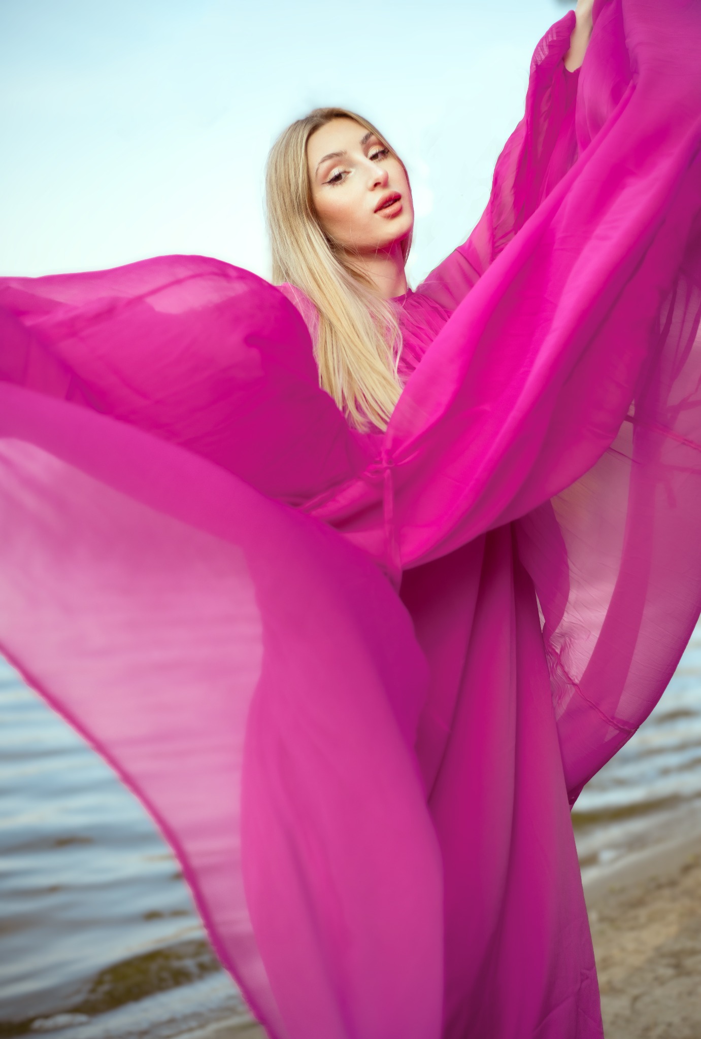 Portret kobiety w zwiewnej, fuksjowej sukni na tle plaży i wody, z lekko rozchylonymi ustami i mocnym makijażem oczu.