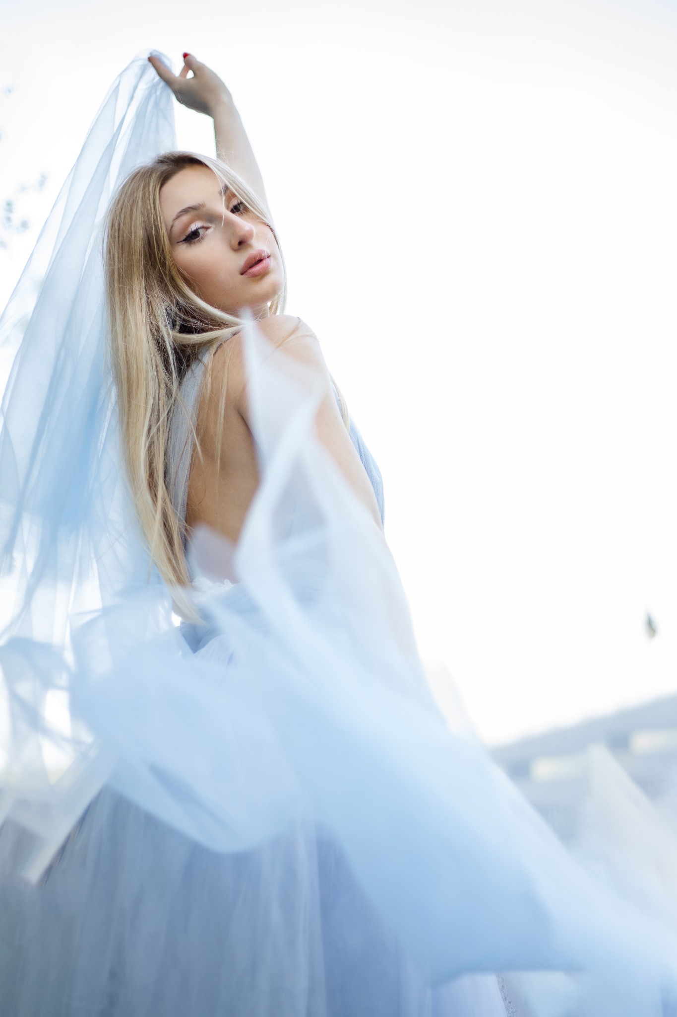 Portret młodej kobiety z blond włosami, trzymającej zwiewny, błękitny materiał, z delikatnym makijażem i czerwonym paznokciem, skierowana lekko w górę, na rozmytym tle.