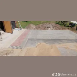 Fragment nowej nawierzchni z kostki brukowej w odcieniach szarości, z elementami czerwonej kostki na obrzeżach, częściowo zasypana piaskiem, widoczny fragment budynku gospodarczego i pryzma ziemi...