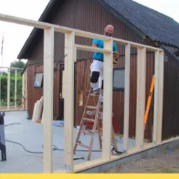 Montaż drewnianej konstrukcji szkieletowej budynku gospodarczego na fundamencie betonowym; pracownik na drabinie z wiertarką, widoczny poziomica i narzędzia.