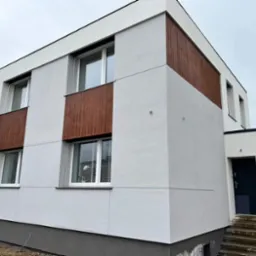 Nowoczesny, dwukondygnacyjny dom z jasnoszarą elewacją i drewnianymi panelami wokół okien, widoczne schody wejściowe i ciemne drzwi.