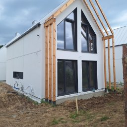 Łukasz Piszczatowski - Nowoczesny dom jednorodzinny z białą elewacją i drewnianą konstrukcją werandy, widoczne czarne okna i przygotowany teren wokół budynku.