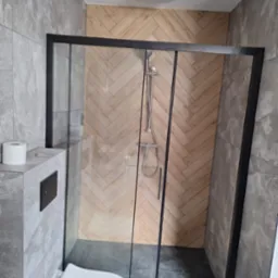 Nowoczesna łazienka z kabiną prysznicową typu walk-in z czarnymi profilami, drewnopodobną ścianą ułożoną w jodełkę i szarą płytką imitującą kamień, widoczny podwieszany sedes z czarnym przyciskiem.