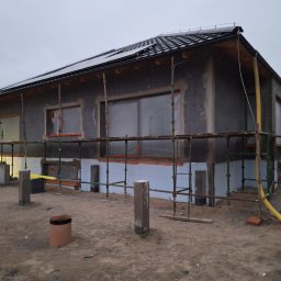 Łukasz Piszczatowski - Dom jednorodzinny w trakcie ocieplania elewacji zewnętrznej, widoczne rusztowania, styropian grafitowy i częściowo zabezpieczone okna folią. Na dachu panele słoneczne.
