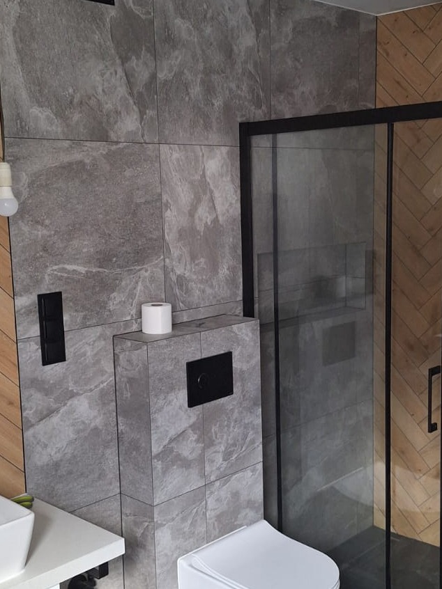 Szara łazienka z kabiną prysznicową typu walk-in, wykończona płytkami imitującymi kamień, z widoczną toaletą podwieszaną i rolką papieru toaletowego na zabudowie.