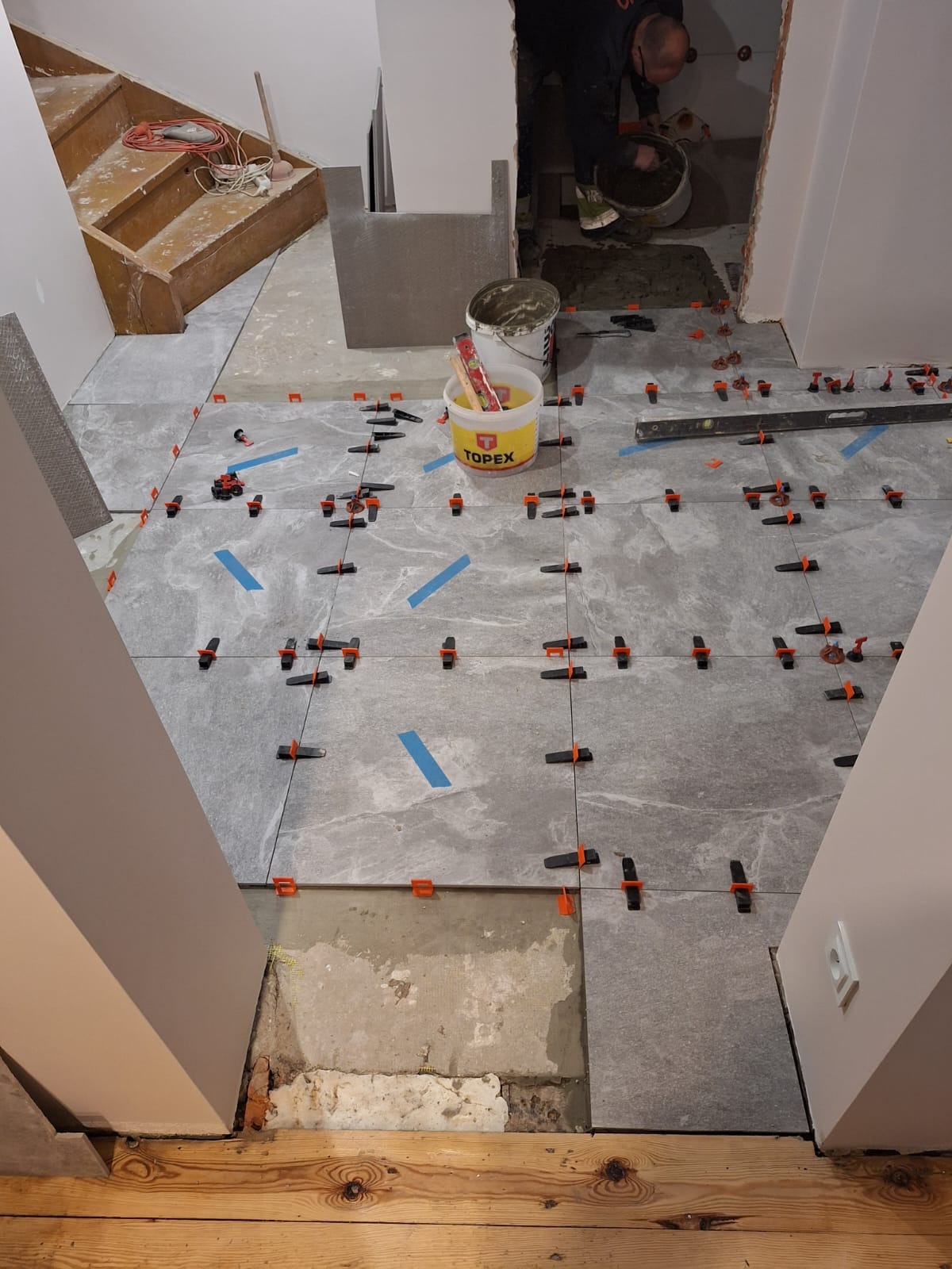 Układanie szarych płytek podłogowych z marmurkowym wzorem, z użyciem klipsów poziomujących i niebieskiej taśmy, widoczny fragment drewnianej podłogi i schody w tle