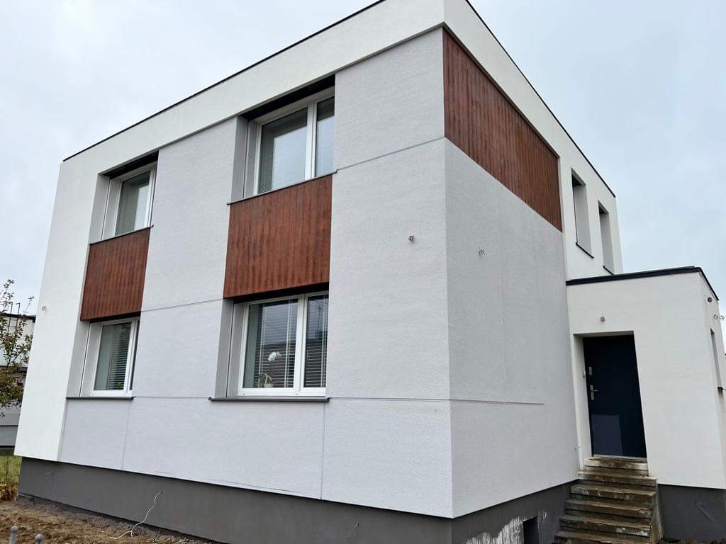 Nowoczesny, dwukondygnacyjny dom z jasnoszarą elewacją i drewnianymi panelami wokół okien, widoczne schody wejściowe i ciemne drzwi.