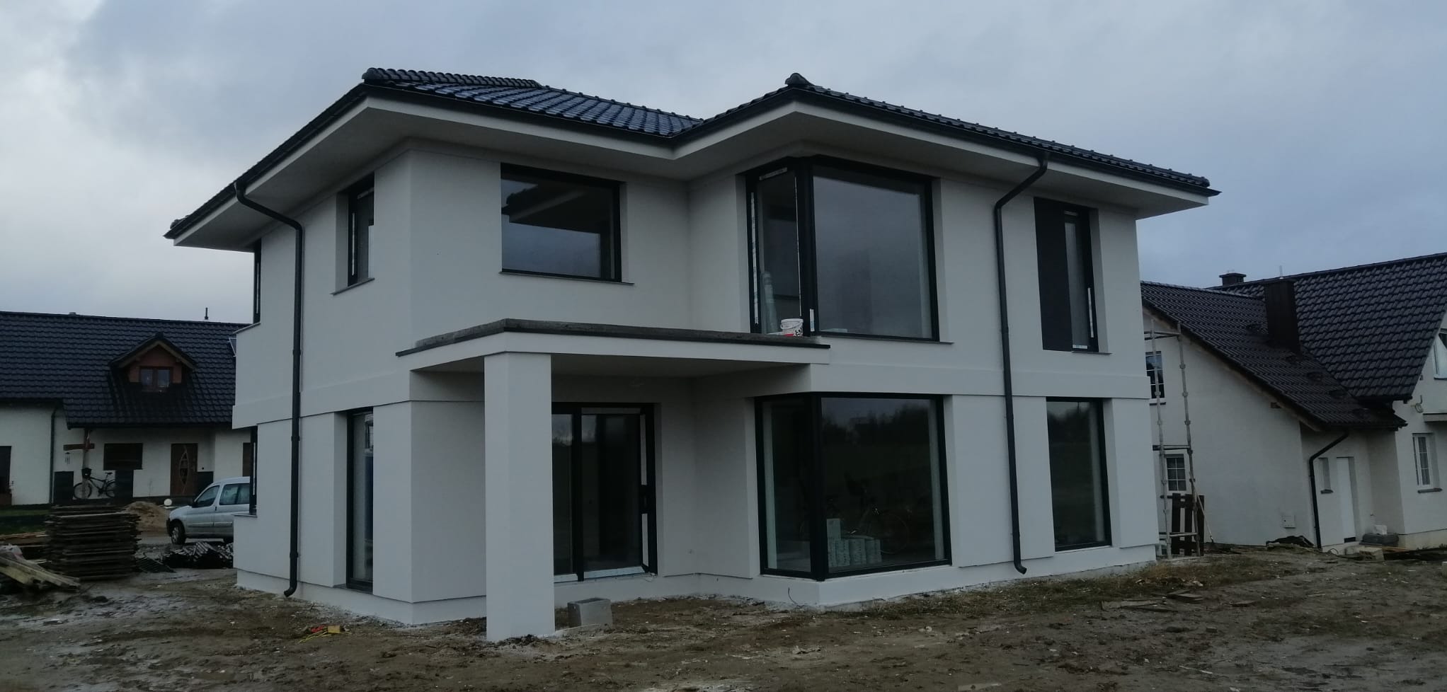 Nowoczesny, dwukondygnacyjny dom z jasną elewacją i czarnymi ramami okiennymi, widoczny z zewnątrz na tle pochmurnego nieba, z widocznym fragmentem sąsiedniego budynku.