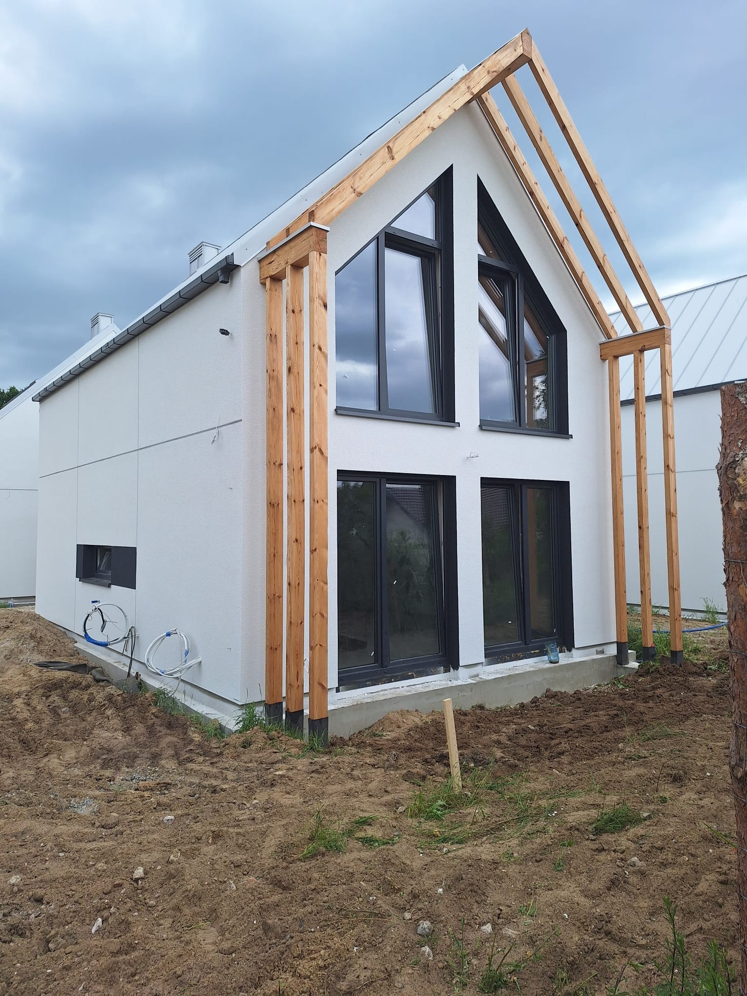 Nowoczesny dom jednorodzinny z białą elewacją i drewnianą konstrukcją werandy, widoczne czarne okna i przygotowany teren wokół budynku.