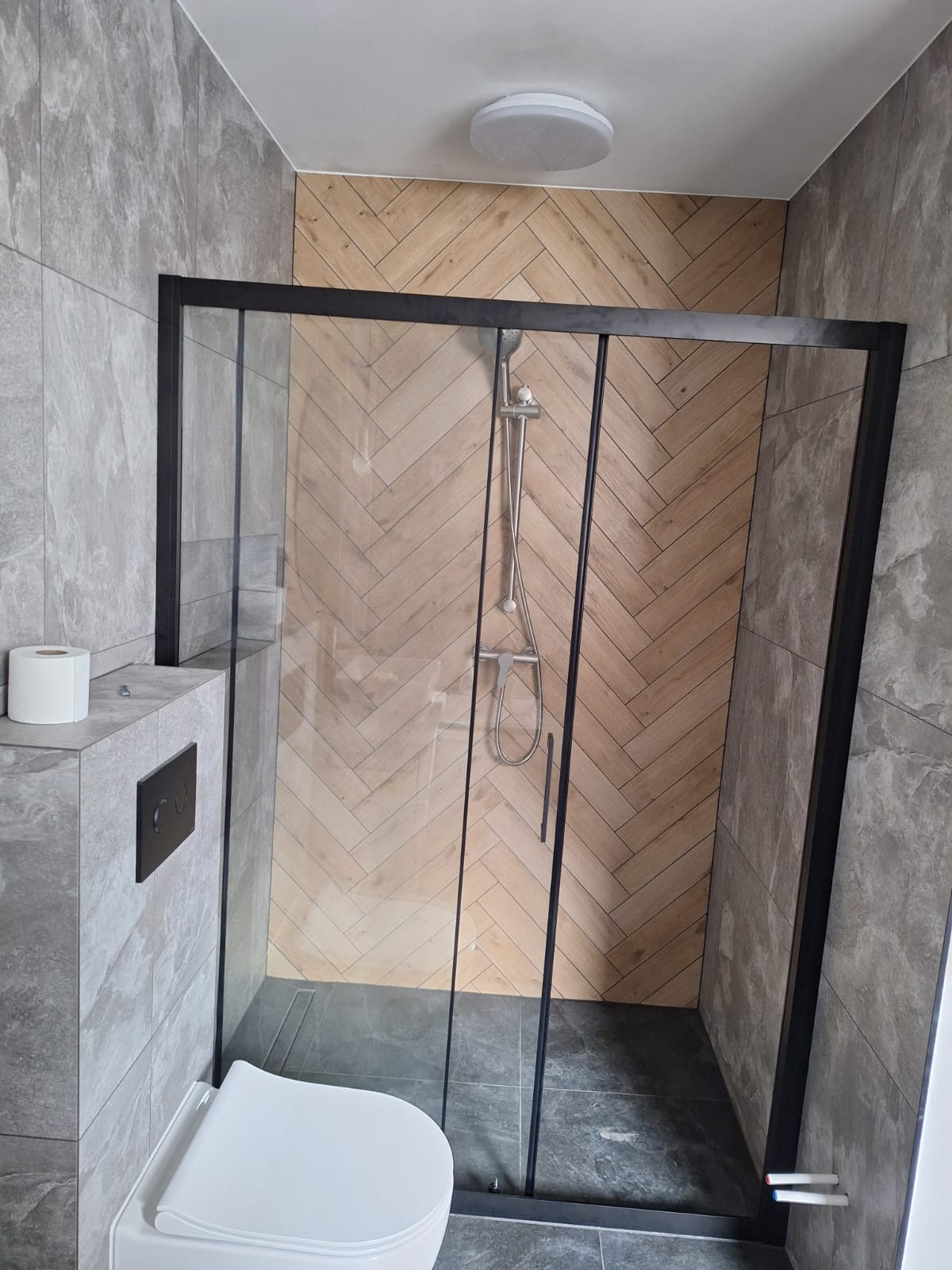 Nowoczesna łazienka z kabiną prysznicową typu walk-in z czarnymi profilami, drewnopodobną ścianą ułożoną w jodełkę i szarą płytką imitującą kamień, widoczny podwieszany sedes z czarnym przyciskiem.