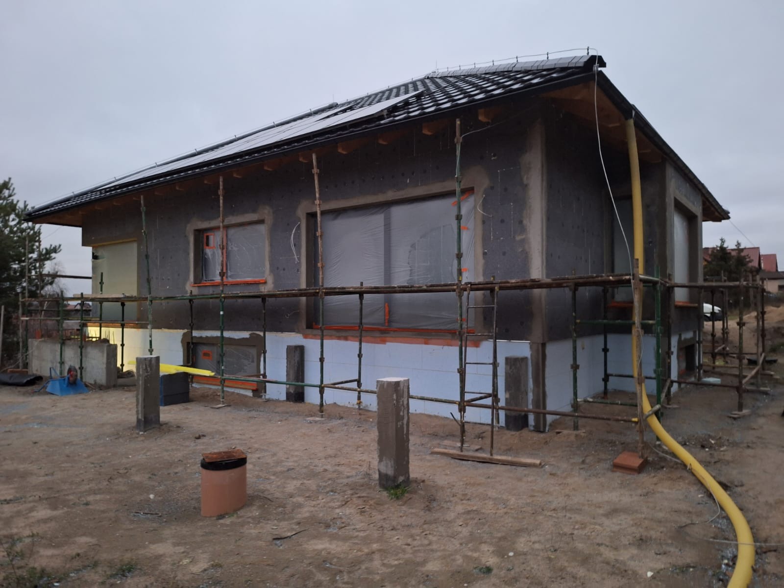 Dom jednorodzinny w trakcie ocieplania elewacji zewnętrznej, widoczne rusztowania, styropian grafitowy i częściowo zabezpieczone okna folią. Na dachu panele słoneczne.