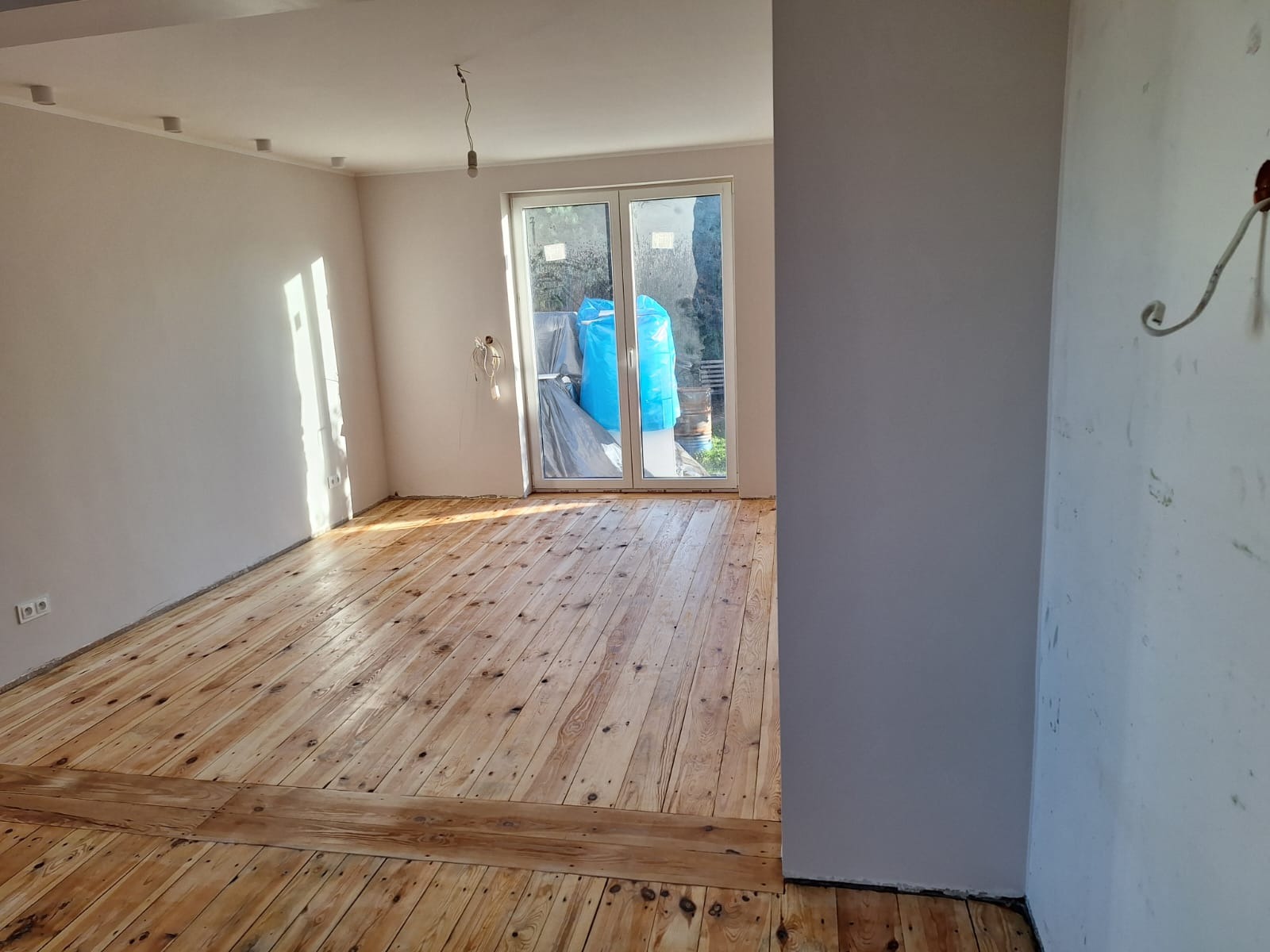 Surowe deski podłogowe w jasnym odcieniu drewna, po renowacji, z wyraźnym usłojeniem, częściowo zabezpieczone listwą przypodłogową, w tle widoczne okno balkonowe z zabezpieczeniem folią malarską...