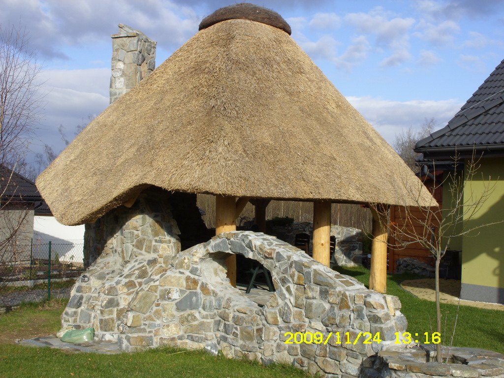 Altana z kamienia i strzechy, z łukowatym wejściem i kamiennym kominem w tle, otoczona zielonym trawnikiem.