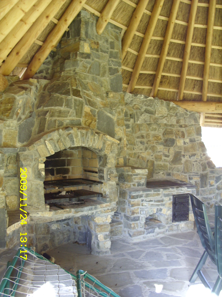 Kamienny grill z kominkiem pod strzechą, z widocznymi rusztami i paleniskiem, otoczony kamienną posadzką i zielonym ogrodzeniem.