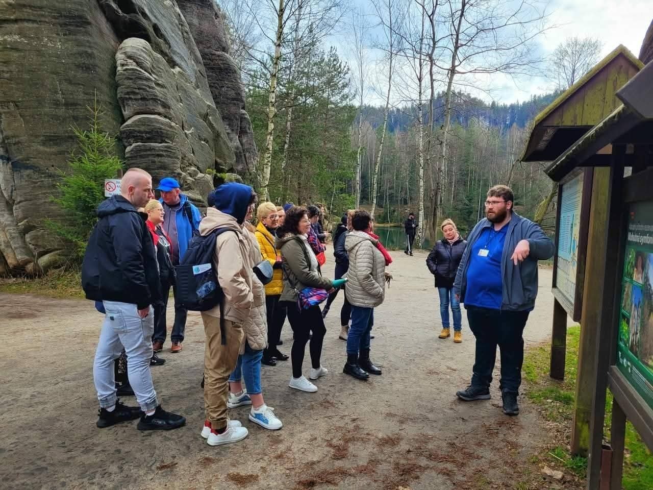 Grupa turystów słucha przewodnika przy tablicy informacyjnej na tle formacji skalnych i lasu.
