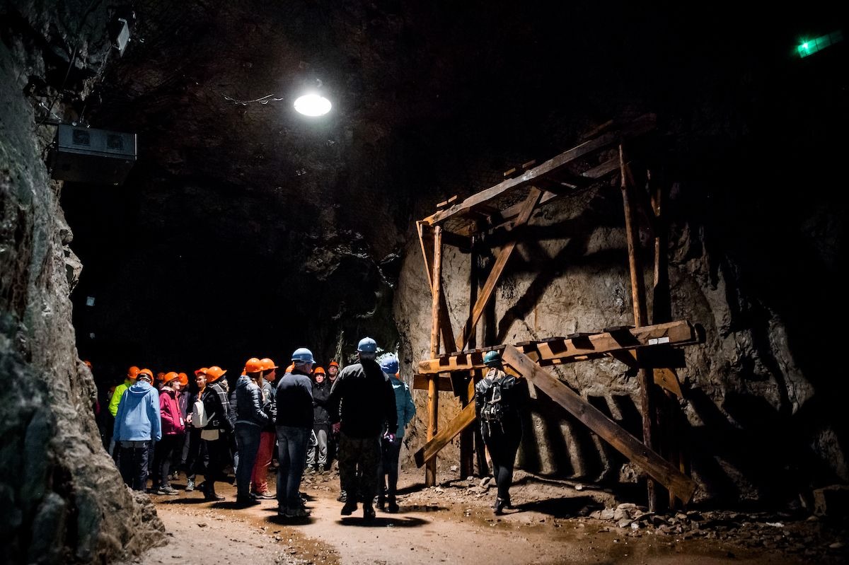 Grupa osób w kaskach zwiedza ciemne wnętrze kopalni z drewnianą konstrukcją zabezpieczającą, oświetloną pojedynczą lampą sufitową.