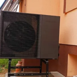 Zewnętrzna jednostka pompy ciepła Samsung na metalowej konstrukcji, zainstalowana przy ścianie budynku z widocznymi panelami słonecznymi w tle.