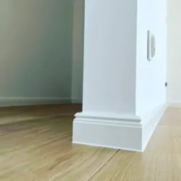 Biała listwa przypodłogowa z frezowanymi detalami, zamontowana na styku ściany i paneli podłogowych o wzorze drewna, widoczny fragment ściany z gniazdkiem.